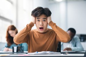 Portrait,Of,Shocked,Asian,Male,Student,Sitting,At,Desk,In