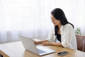 Woman,Taking,Notes,While,Working,On,Laptop,At,Desk